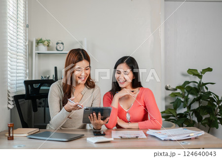 Two women working together on a tablet in a bright office, surrounded by documents and plants, showcasing teamwork and modern technology. 124640043