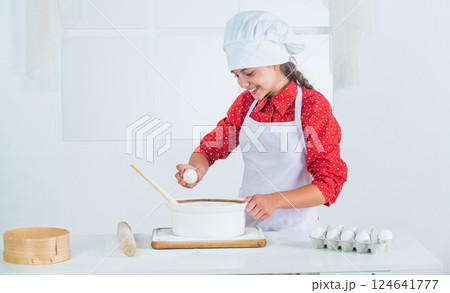 adding the egg. time to eat. happy child cooking in kitchen. bake cookies in kitchen. professional and skilled baker. kid in chef uniform and hat. teen girl preparing dough. making cake by recipe 124641777