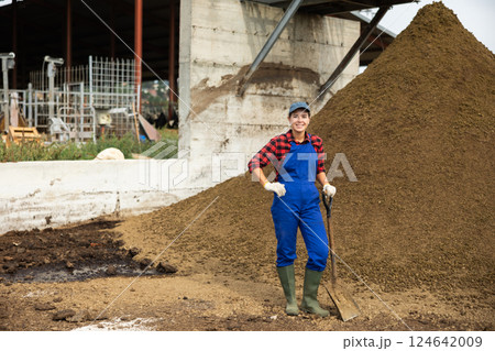 Female farmer with shovel poses against pile of dried and crushed cow dung in the backyard of farm 124642009