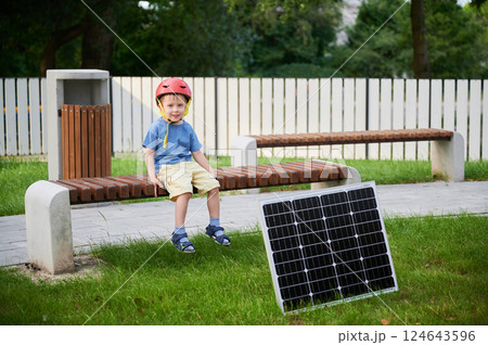 Young boy sits on wooden bench in park, smiling at the camera, wearing red helmet. Beside on grass is solar panel. Concept of green energy generation. Young boy sits on wooden bench in park, smiling at the camera, wearing red helmet. Beside on grass is solar panel. Concept of green energy generation. 124643596