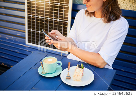 Young woman charging her smartphone from solar panel while relaxing. Pretty girl drinking coffee and checking her phone next to solar battery. Conscious female choosing sustainable lifestyle. Young woman charging her smartphone from solar panel while relaxing. Pretty girl drinking coffee and checking her phone next to solar battery. Conscious female choosing sustainable lifestyle. 124643625