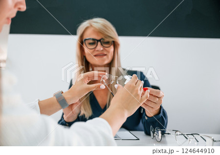 Sitting by the table. Woman choosing glasses in the store and getting help by assistant Sitting by the table. Woman choosing glasses in the store and getting help by assistant 124644910