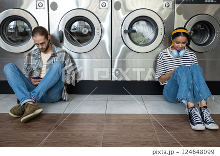 Man and woman in laundromat waiting for washing machines while scrolling online on smartphones 124648099