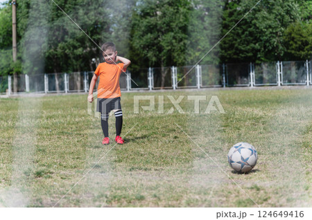 Little boy in orange t-shirt stands on grassy field in front of soccer ball, seen through goal net. 124649416