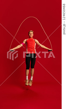 Fit young man in red training top jumping with skipping rope mid-air, showing focus and agility in active motion against red background. Fit young man in red training top jumping with skipping rope mid-air, showing focus and agility in active motion against red background. 124650468