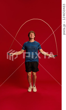 Dynamic photo of young man doing high-speed jump rope workout against red background, expressing joy and movement. 124650483
