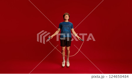 Energetic man mid-air skipping rope, smiling during dynamic cardio session against red studio background. Energetic man mid-air skipping rope, smiling during dynamic cardio session against red studio background. 124650484