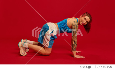 Woman in blue sports shirt and black shorts standing on all fours, smiling confidently, preparing for workout. 124650490