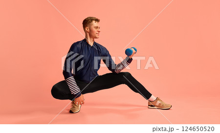 Sporty male in wide lunge holding dumbbell in bicep curl, wearing blue athletic outfit, smiling during workout on pink background. Sporty male in wide lunge holding dumbbell in bicep curl, wearing blue athletic outfit, smiling during workout on pink background. 124650520