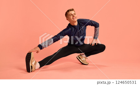 Sporty man smiling while stretching legs sideways, reaching for foot on pink background, wearing striped sleeve sportswear. Sporty man smiling while stretching legs sideways, reaching for foot on pink background, wearing striped sleeve sportswear. 124650531