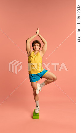 Smiling sporty man in bright gym outfit standing in tree pose on foam block, practicing balance and focus against pink background. 124650538