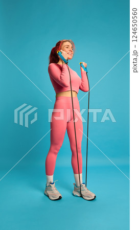 Young woman in pink activewear exercising with resistance band, doing bicep curls, showing muscle engagement and positive attitude. Young woman in pink activewear exercising with resistance band, doing bicep curls, showing muscle engagement and positive attitude. 124650560