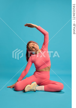 Young sporty woman stretching sideways on floor in pink activewear, smiling and showing flexibility against blue studio background. Young sporty woman stretching sideways on floor in pink activewear, smiling and showing flexibility against blue studio background. 124650561