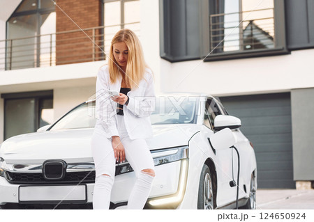With smartphone. Young woman in white clothes is with her electric car at daytime With smartphone. Young woman in white clothes is with her electric car at daytime 124650924