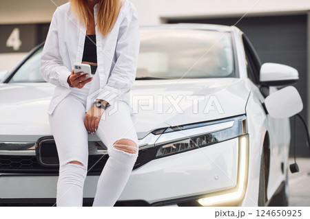 With smartphone. Young woman in white clothes is with her electric car at daytime 124650925