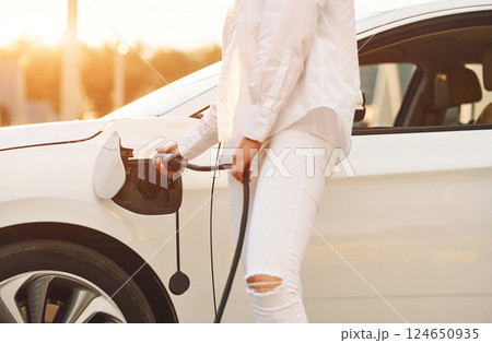 Holding charger. Young woman in white clothes is with her electric car at daytime 124650935