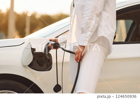 Holding charger. Young woman in white clothes is with her electric car at daytime 124650936