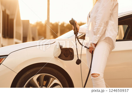 Holding charger. Young woman in white clothes is with her electric car at daytime 124650937