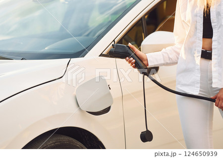 Close up view. Holding charger. Young woman in white clothes is with her electric car at daytime 124650939