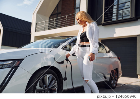 Charging process. Young woman in white clothes is with her electric car at daytime Charging process. Young woman in white clothes is with her electric car at daytime 124650940