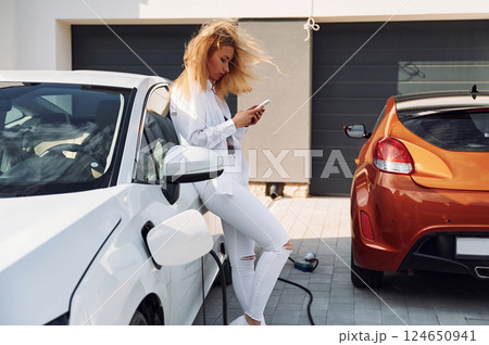 Charging process. Young woman in white clothes is with her electric car at daytime 124650941