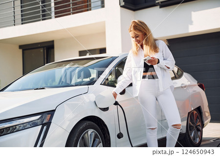 Charging process. Young woman in white clothes is with her electric car at daytime 124650943