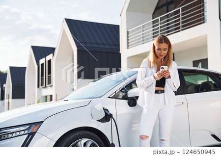 Waiting automobile to charge. Young woman in white clothes is with her electric car at daytime 124650945