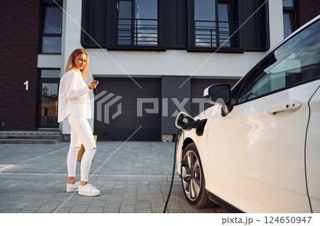 Waiting automobile to charge. Young woman in white clothes is with her electric car at daytime 124650947