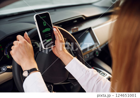 Sitting inside with smartphone. Young woman in white clothes is with her electric car at daytime 124651001