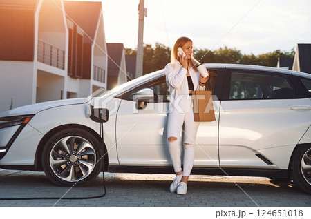 Holding shopping bag. Young woman in white clothes is with her electric car at daytime 124651018