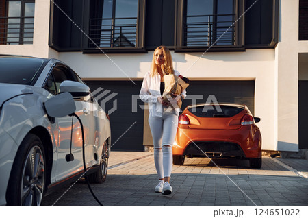 Holding shopping bag. Young woman in white clothes is with her electric car at daytime Holding shopping bag. Young woman in white clothes is with her electric car at daytime 124651022