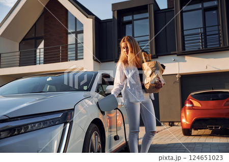Holding shopping bag. Young woman in white clothes is with her electric car at daytime Holding shopping bag. Young woman in white clothes is with her electric car at daytime 124651023
