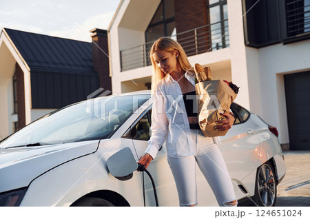 Holding shopping bag. Young woman in white clothes is with her electric car at daytime Holding shopping bag. Young woman in white clothes is with her electric car at daytime 124651024
