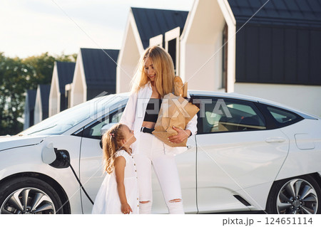 Standing together. Young woman with her little daughter is with their electric car outdoors Standing together. Young woman with her little daughter is with their electric car outdoors 124651114
