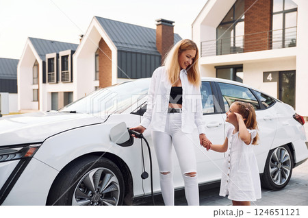 Standing together. Young woman with her little daughter is with their electric car outdoors 124651121