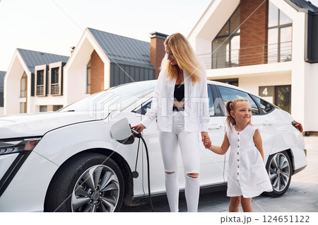 Standing together. Young woman with her little daughter is with their electric car outdoors 124651122