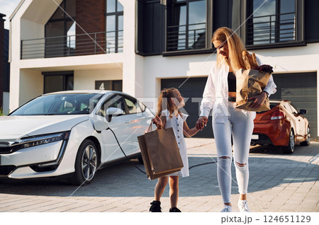 Holding bags while vehicle is on a charge. Young woman with her little daughter is with their electric car outdoors 124651129