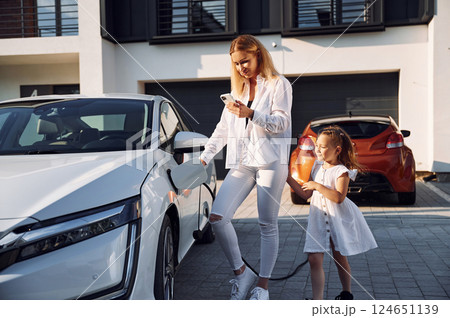 Standing at daytime together. Waiting for a charge. Young woman with her little daughter is with their electric car outdoors 124651139