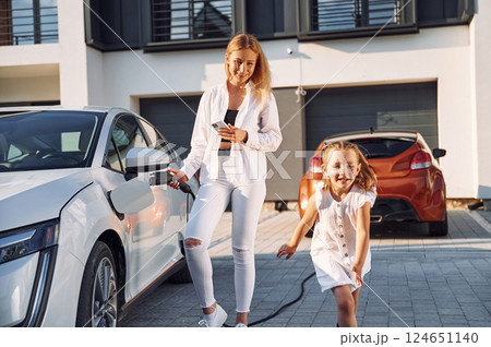 Standing at daytime together. Waiting for a charge. Young woman with her little daughter is with their electric car outdoors 124651140