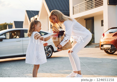 Walking and holding bags with products. Young woman with her little daughter is with their electric car outdoors Walking and holding bags with products. Young woman with her little daughter is with their electric car outdoors 124651158