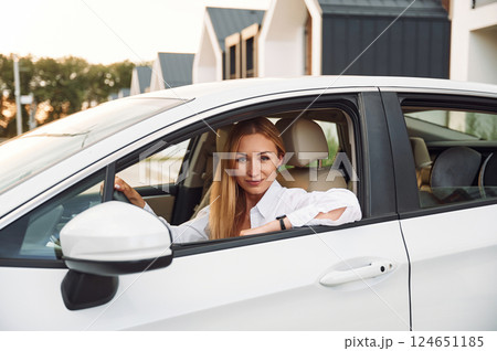 Sitting and looking at the camera. Young woman in white clothes is with her electric car at daytime 124651185