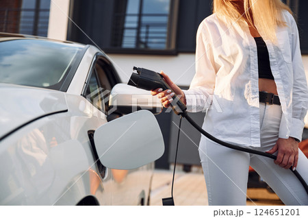 Putting charging cable into the vehicle. Young woman in white clothes is with her electric car at daytime 124651201