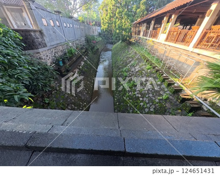 A narrow canal runs between buildings with stone walls. Lush greenery and trees are visible. A narrow canal runs between buildings with stone walls. Lush greenery and trees are visible. 124651431