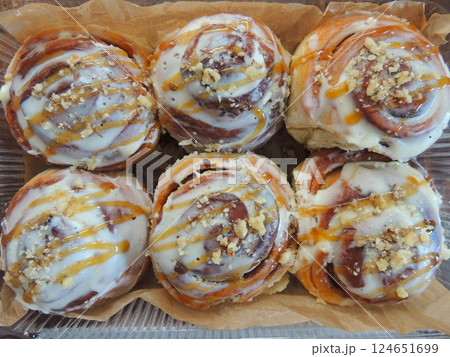 six homemade twisted buns with glaze and nut sprinkles lying on brown parchment paper, freshly baked cinnabons close-up with sweet white filling and nut crumbs 124651699