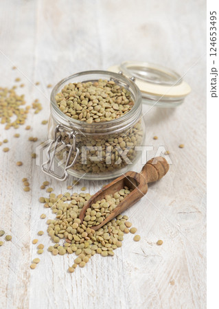 Glass jar full of raw green lentils with a wooden scoop on white table closeup. Dried legumes 124653495
