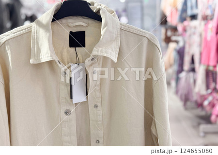A beige button-up shirt with a blank price tag hangs on a rack in a brightly lit clothing store 124655080
