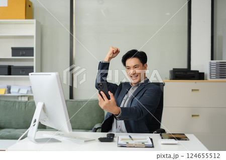 Successful business man, excited young man Keeping up arms and showing positive emotions, happy working in modern office on smartphone. 124655512
