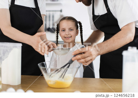 Caucasian family with young girl cooking in bright kitchen. Focus on teaching and teamwork. Includes flour, bowl, whisk engaging girl with smiling face and braided hair. Caucasian family with young girl cooking in bright kitchen. Focus on teaching and teamwork. Includes flour, bowl, whisk engaging girl with smiling face and braided hair. 124656218