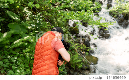 Man wearing an orange vest holds a black cat next to a cascading waterfall in lush forest. Traveling with pet, walking and adventurous , serene outdoor moment amidst nature Man wearing an orange vest holds a black cat next to a cascading waterfall in lush forest. Traveling with pet, walking and adventurous , serene outdoor moment amidst nature 124656469