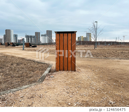 Wooden trash can in the foreground of an urban park, pathways and barren trees visible. High-rise buildings loom in the background, under a cloudy sky, urban development copy space 124656475